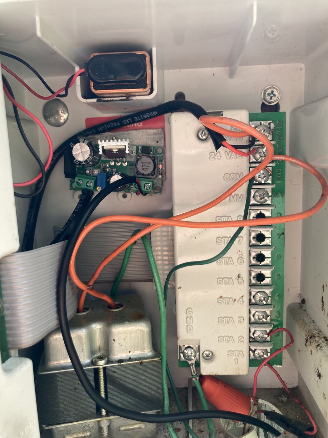 Close-up of the 24VAC terminal block and a small adjustable buck module marked '470' with a trimpot. Orange wire jumps from the 24VAC terminal to the module input; output pair leaves through the cable gland to the Node.