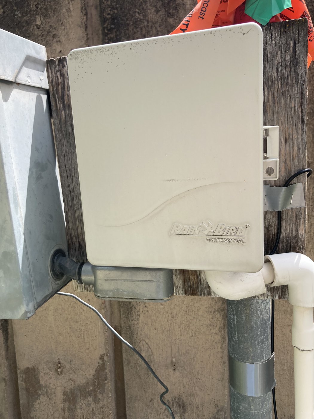 Rain Bird Professional irrigation controller mounted on a weathered wood post, lid closed, with a metal conduit gland and PVC drop visible at the bottom. Comcast utility flag in the foreground.