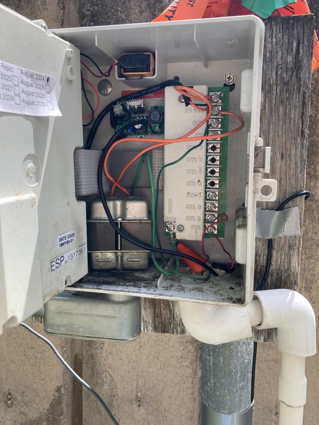 Face panel swung out, revealing the controller's internal terminal strip with screw terminals for 24VAC, common, master valve, and station outputs. Existing irrigation wires routed in, with a small green buck-converter PCB tucked into the lower left, taped in place.