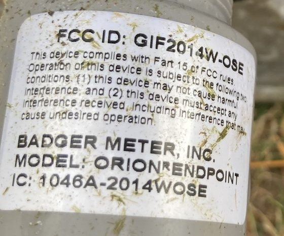 A cylindrical ORION water-meter endpoint lifted out of the well, with its FCC label facing the camera: 'FCC ID: GIF2014W-OSE', 'BADGER METER, INC.', 'MODEL: ORION ENDPOINT', 'IC: 1046A-2014WOSE'.