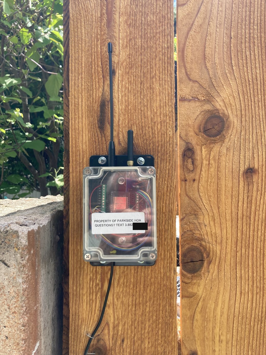 Close-up of the deployed Node enclosure: a clear-lidded plastic project box with two short whip antennas on the left side, an HOA-property identification label visible through the lid, and an external cable exiting the right side.