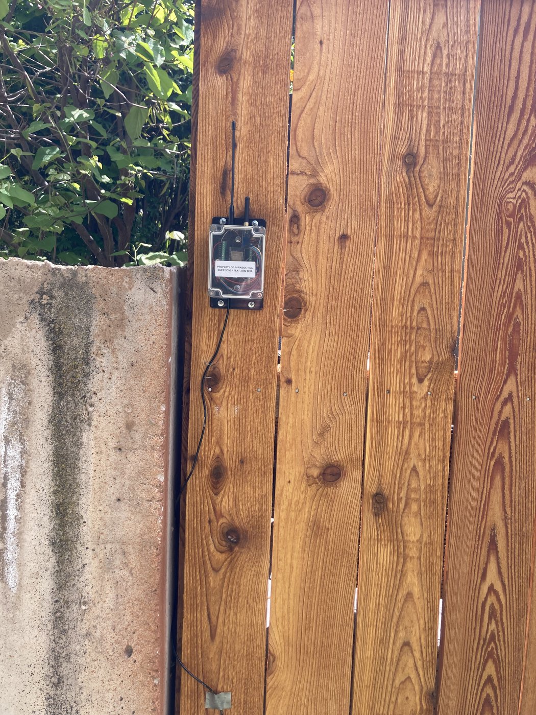 Closer view of the same cedar wall, the mounted device now clearly visible: a small black-bezeled box screwed to one of the lower cedar boards, with a thin power cable running out of frame to the right.