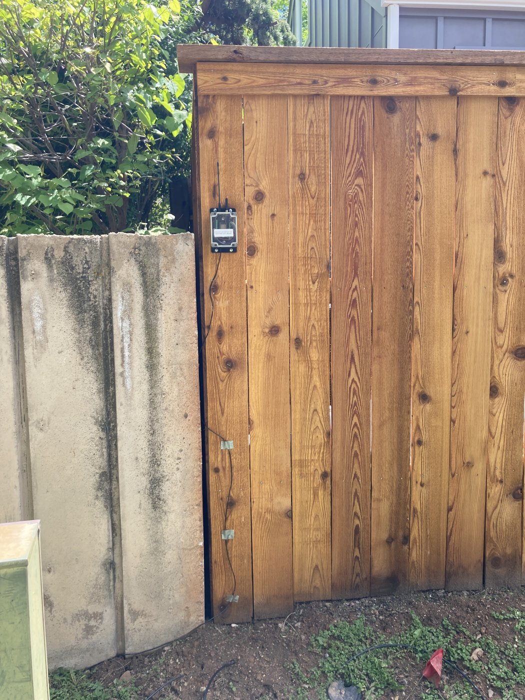 Wide view of a cedar-board exterior wall meeting a concrete walkway, with a small dark device mounted near the bottom edge of the boards and a shrub at the right. The mount sits low, inconspicuous, and out of the homeowner's traffic path.