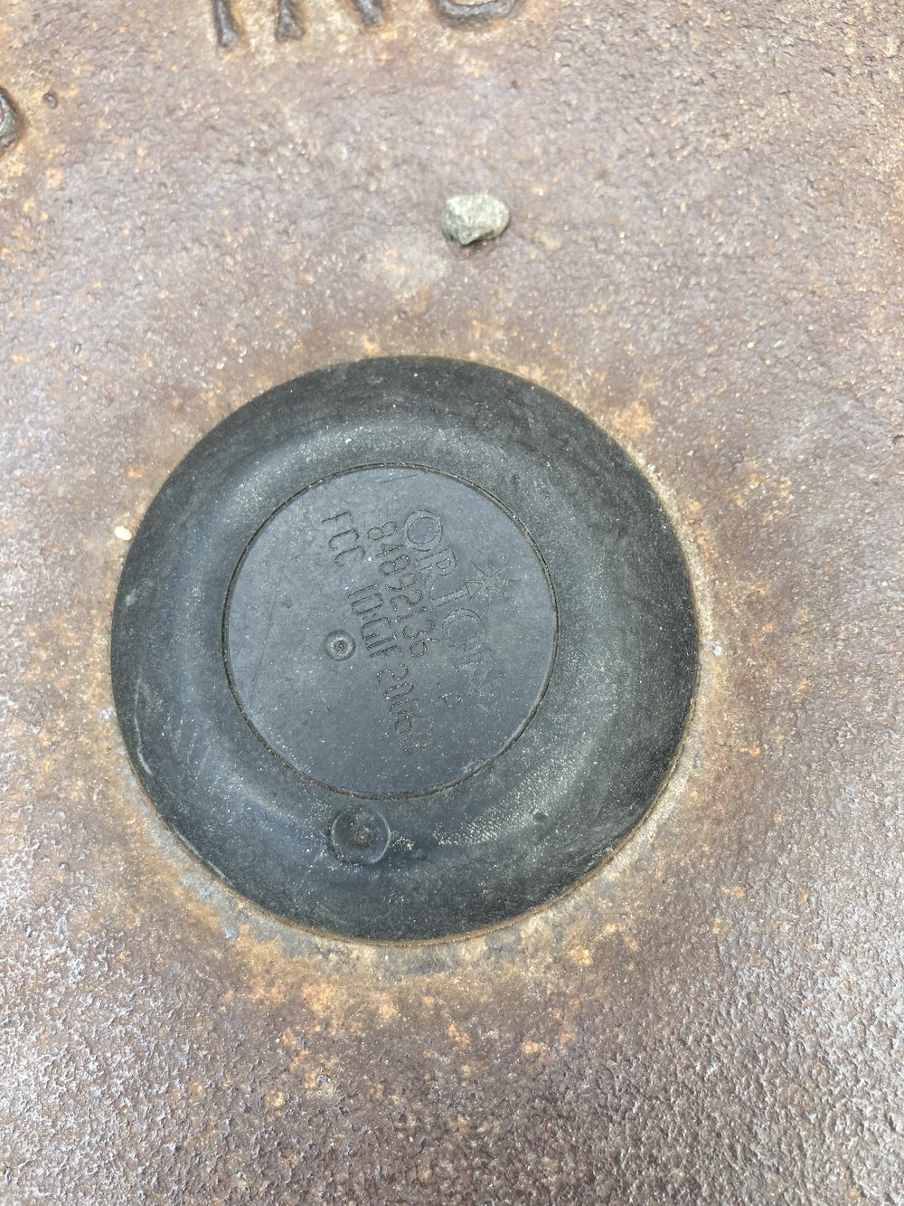 A water meter well cover viewed from above, with a round black plastic ORION radio endpoint disk seated in a recess at its center, molded with an ORION logo and an FCC ID. This is the part the Node receives over 900 MHz.